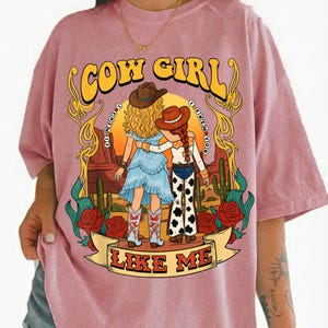 May include: A dusty pink t-shirt featuring a graphic print of two figures in cowboy hats standing in a desert landscape with cacti and red roses. The text reads COW GIRL at the top and LIKE ME on a banner at the bottom, with I KNEW IT I KNEW YOU.