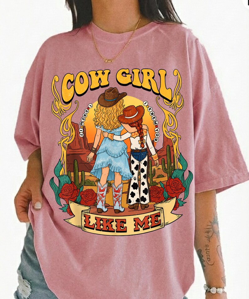 May include: A dusty pink t-shirt featuring a graphic print of two figures in cowboy hats standing in a desert landscape with cacti and red roses. The text reads COW GIRL at the top and LIKE ME on a banner at the bottom, with I KNEW IT I KNEW YOU.