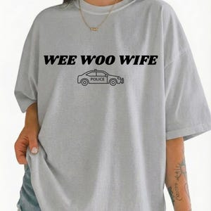 Può includere: T-shirt grigio chiaro con la scritta nera "WEE WOO WIFE" sopra una grafica di un'auto della polizia in bianco e nero. La maglietta ha maniche corte e una vestibilità ampia.