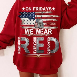 Può includere: Una felpa rossa con la scritta "ON FRIDAYS WE WEAR RED" e un disegno della bandiera americana. Il disegno include targhette militari. Le parole "REMEMBER EVERYONE DEPLOYED" sono anch'esse stampate.