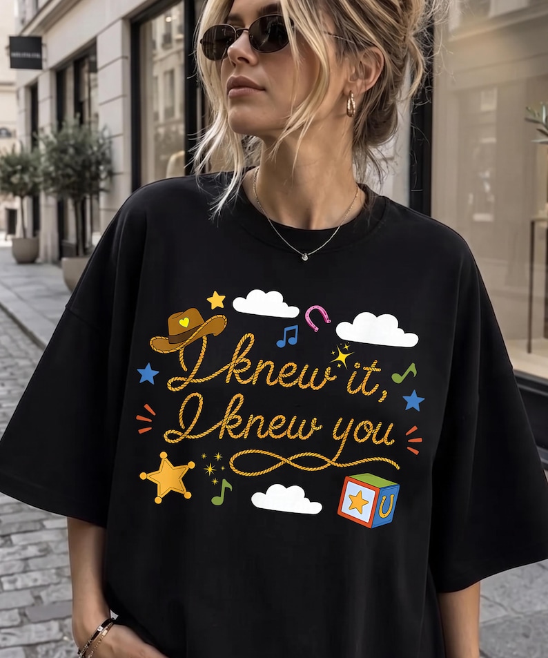 May include: A black t-shirt featuring a graphic print with the text "I knew it, I knew you" in a rope-style font. The design includes a brown cowboy hat, white clouds, a yellow star, a colorful block, musical notes, and a pink horseshoe.