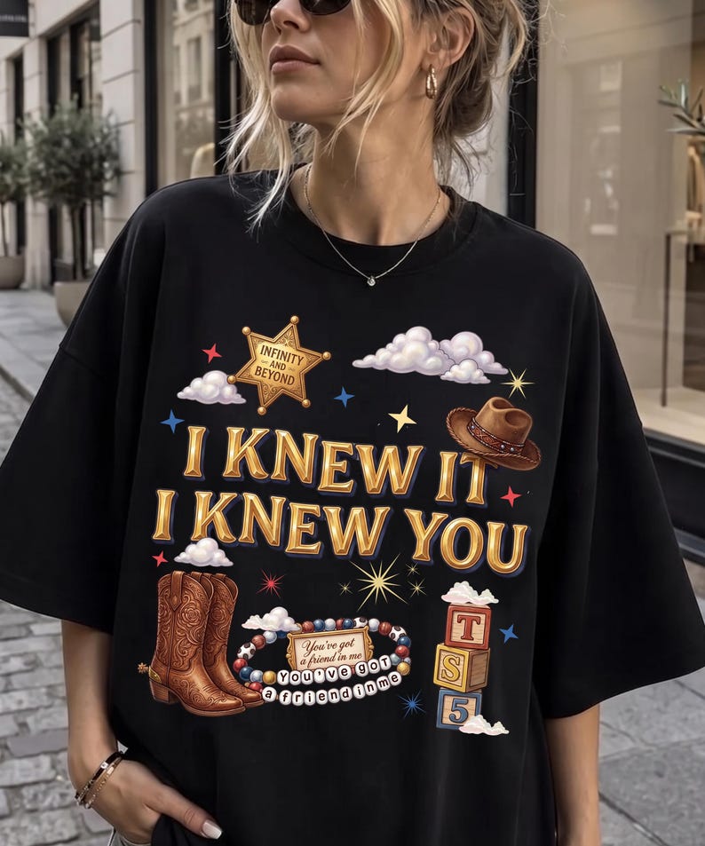 May include: A black short-sleeved t-shirt featuring a graphic print with the text "I KNEW IT I KNEW YOU". The design includes a gold sheriff star labelled "INFINITY AND BEYOND", a brown cowboy hat, brown cowboy boots, a beaded bracelet, and blocks.