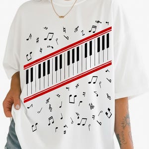 Könnte beinhalten: Weißes T-Shirt mit einem Klaviertasten-Design. Die Tastatur hat schwarz-weiße Tasten, die von roten Streifen umrahmt sind. Schwarze Noten sind um die Tastatur herum verstreut. Ein lässiges Oberteil zum Thema Musik.