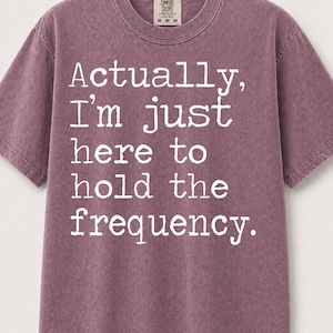 Op de afbeelding: Een mauve Comfort Colors T-shirt met de witte tekst "Actually, I'm just here to hold the frequency". Het shirt heeft een ronde hals en korte mouwen. Het label luidt M-M-M.