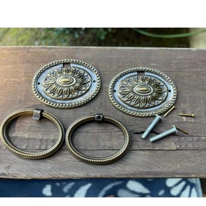 May include: Two oval antique brass drawer pulls featuring a central floral motif with a beaded border. The set includes two matching oval backplates, two circular ring handles, and three small metal screws for installation and mounting.