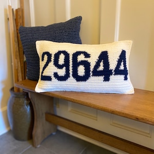 May include: Two crochet pillows on a wooden bench. One is navy blue, the other is cream-colored with the number "29644" in navy blue. The pillows are decorative and add a touch of handmade charm to any space.