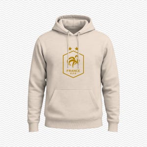 Puede incluir: Una sudadera con capucha de color beige que presenta un gr&aacute;fico de color dorado en el pecho. El gr&aacute;fico muestra un contorno hexagonal con un emblema de gallo, dos estrellas encima y el texto FRANCE FFF en letras doradas debajo.