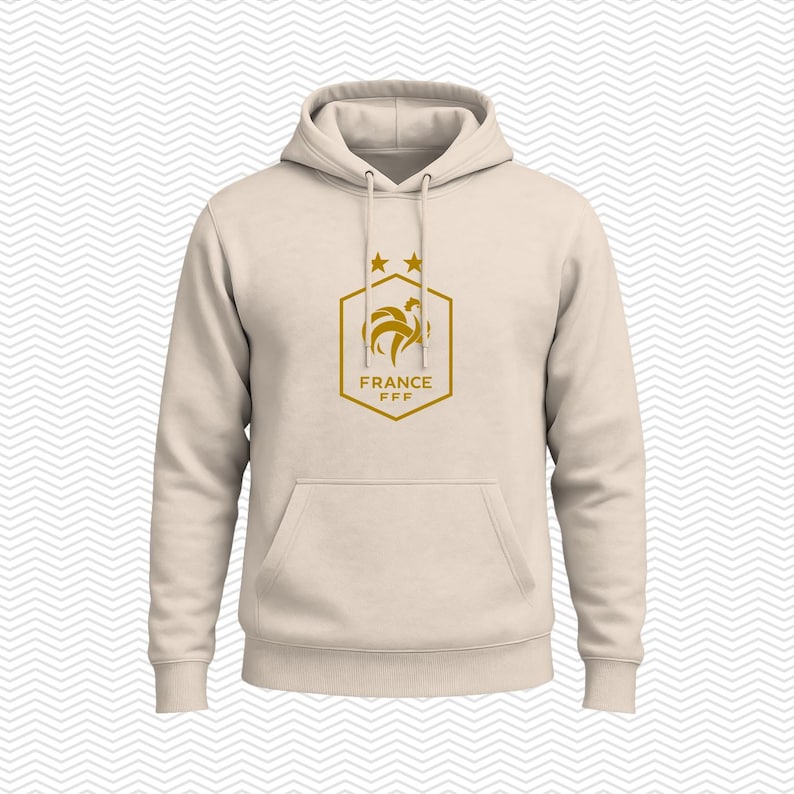 Puede incluir: Una sudadera con capucha de color beige que presenta un gr&aacute;fico de color dorado en el pecho. El gr&aacute;fico muestra un contorno hexagonal con un emblema de gallo, dos estrellas encima y el texto FRANCE FFF en letras doradas debajo.