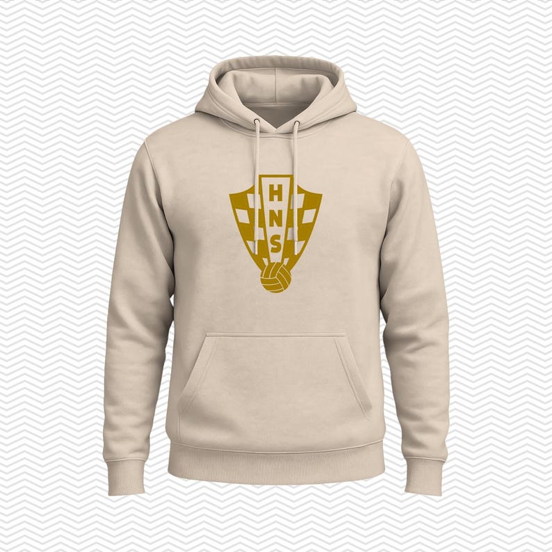 May include: A beige hooded sweatshirt featuring a gold-colored graphic on the chest. The graphic displays a shield with a checkered pattern, the letters HNS stacked vertically, and a soccer ball icon at the bottom. The hoodie has long sleeves.