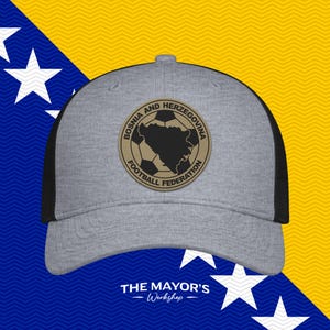 May include: A grey baseball cap with black mesh back panels. The front features a circular tan patch with a black map of Bosnia and Herzegovina, a soccer ball graphic, and the text "BOSNIA AND HERZEGOVINA FOOTBALL FEDERATION" in black.