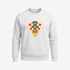 May include: A white long-sleeve sweatshirt featuring a centered graphic of the Croatian Football Federation crest. The shield-shaped emblem displays a red and white checkerboard pattern, a blue vertical stripe with the yellow letters HNS, and a soccer ball.