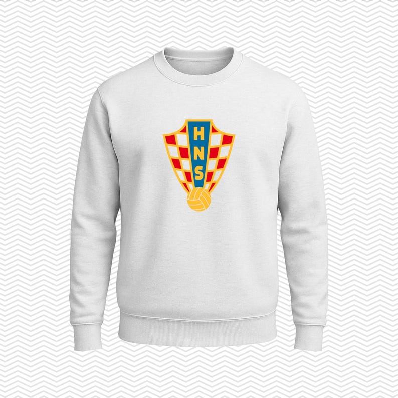 May include: A white long-sleeve sweatshirt featuring a centered graphic of the Croatian Football Federation crest. The shield-shaped emblem displays a red and white checkerboard pattern, a blue vertical stripe with the yellow letters HNS, and a soccer ball.