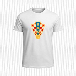 May include: A white short-sleeved t-shirt featuring a centered graphic of the Croatian Football Federation crest. The shield-shaped emblem displays a red and white checkered pattern, a blue vertical stripe with the letters HNS, and a yellow ball.