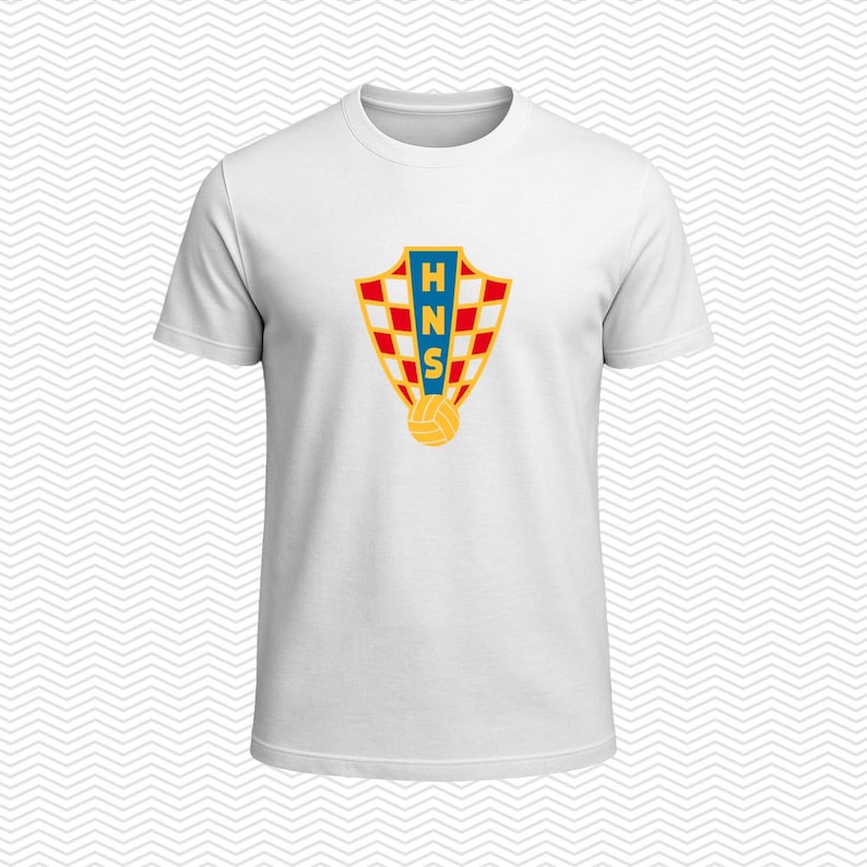May include: A white short-sleeved t-shirt featuring a centered graphic of the Croatian Football Federation crest. The shield-shaped emblem displays a red and white checkered pattern, a blue vertical stripe with the letters HNS, and a yellow ball.
