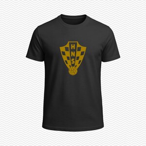 May include: A black short-sleeved t-shirt featuring a gold-colored graphic on the chest. The graphic displays a shield with a checkered pattern, the letters HNS vertically aligned in the center, and a small soccer ball icon positioned below the shield.