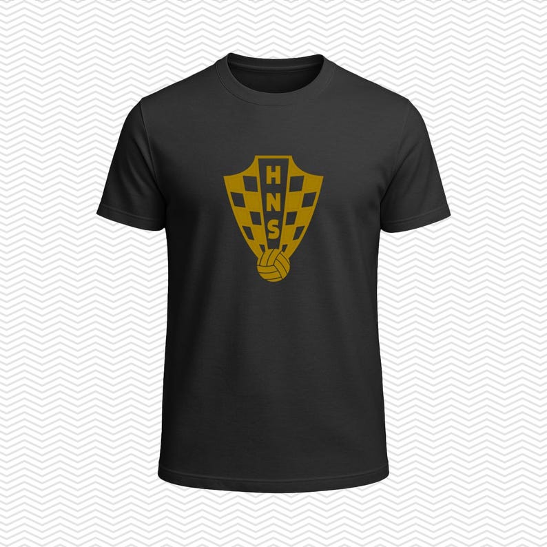 May include: A black short-sleeved t-shirt featuring a gold-colored graphic on the chest. The graphic displays a shield with a checkered pattern, the letters HNS vertically aligned in the center, and a small soccer ball icon positioned below the shield.