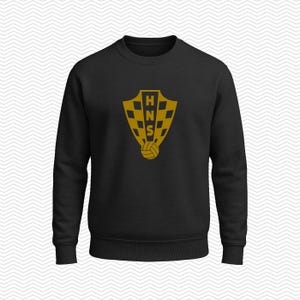 May include: A black long-sleeve crewneck sweatshirt featuring a gold-colored graphic on the chest. The graphic displays a shield with a checkered pattern, the letters HNS stacked vertically in the center, and a soccer ball at the bottom.