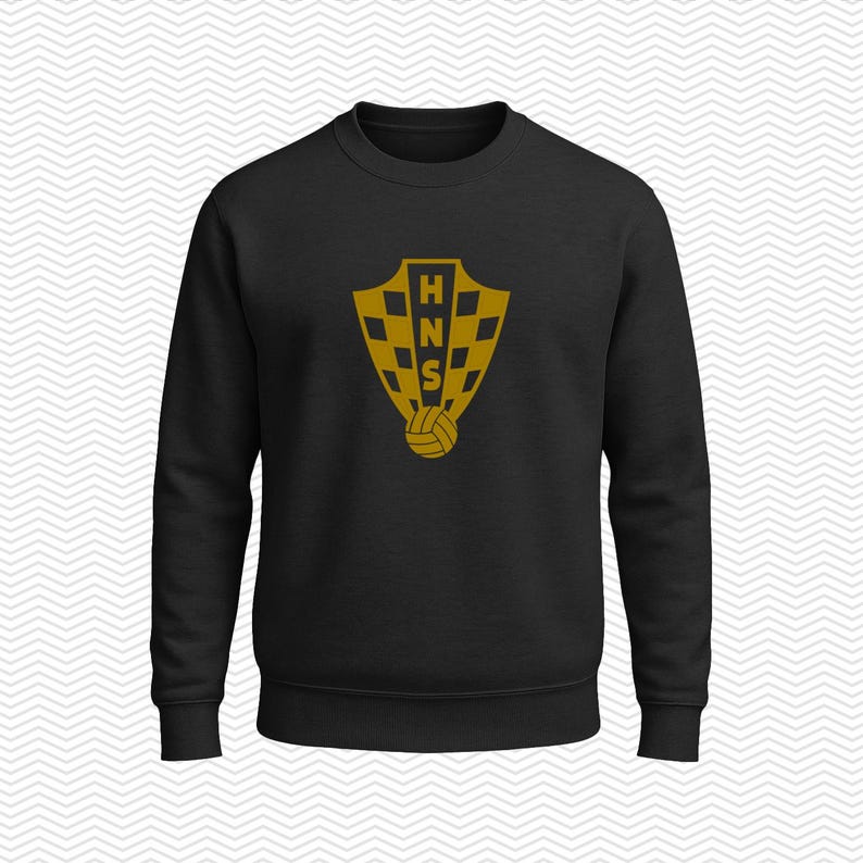 May include: A black long-sleeve crewneck sweatshirt featuring a gold-colored graphic on the chest. The graphic displays a shield with a checkered pattern, the letters HNS stacked vertically in the center, and a soccer ball at the bottom.