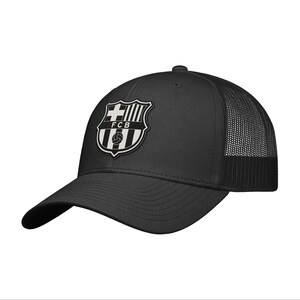 May include: A black baseball cap featuring a mesh back and a structured front panel. The front displays a black and white embroidered patch of the FCB crest, which includes a cross, vertical stripes, and a soccer ball graphic with the text FCB.