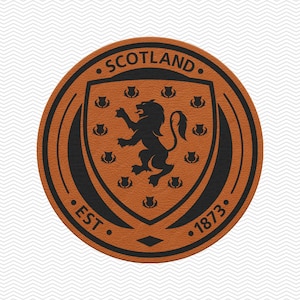 May include: A round, brown leather patch featuring the Scotland coat of arms. The design includes a black lion rampant, thistles, and the text "SCOTLAND" and "EST 1873". The patch has a textured appearance.