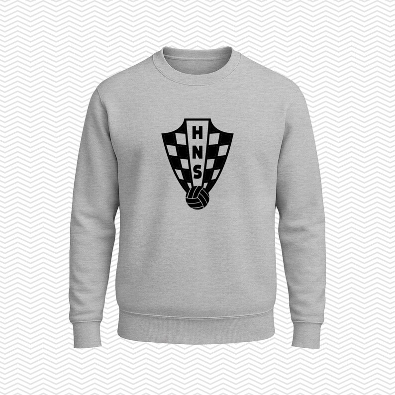 May include: A light gray crewneck sweatshirt featuring a black graphic on the chest. The graphic displays a shield with a checkered pattern, the letters HNS stacked vertically in the center, and a soccer ball positioned at the base of the shield.