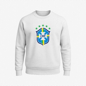 May include: A white long-sleeve crewneck sweatshirt featuring the blue, green, and yellow CBF crest logo centered on the chest. The logo includes a shield shape with a cross, vertical stripes, and five green stars arranged in an arc above it.
