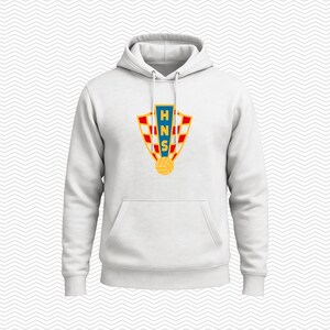 May include: A white hooded sweatshirt featuring a central graphic of a shield with a red and white checkered pattern. The shield contains a blue vertical stripe with the yellow letters HNS and a yellow soccer ball icon positioned at the base.