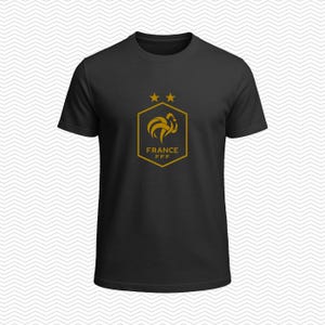 Puede incluir: Una camiseta negra de manga corta con un gr&aacute;fico de color dorado en el pecho. El dise&ntilde;o incluye un emblema hexagonal con un gallo estilizado, dos estrellas sobre el hex&aacute;gono y el texto FRANCE FFF impreso dentro del emblema.