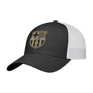 May include: A black baseball cap with a white mesh back and a tan-colored patch on the front featuring the FCB crest. The cap has a curved brim and a structured front panel, displaying the shield emblem with a cross, stripes, and a soccer ball.