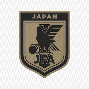 May include: A shield-shaped patch featuring the Japanese national team emblem. The top of the patch reads "JAPAN" in white letters. Below, a black bird with outstretched wings and a ball is centered above the letters "JFA". The patch has a textured, tan background.