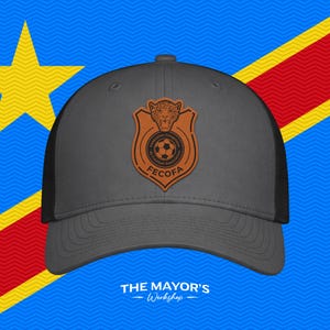 May include: A charcoal gray baseball cap with a black mesh back and a brown leather-like patch on the front. The patch features a leopard head above a soccer ball and the text FECOFA and Fédération Congolaise de Football Association.