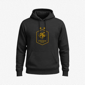 Puede incluir: Una sudadera con capucha negra que presenta un gr&aacute;fico de color dorado en el pecho. El gr&aacute;fico muestra un emblema hexagonal con la silueta de un gallo, dos estrellas encima y el texto FRANCE FFF impreso en letras doradas dentro del hex&aacute;gono.
