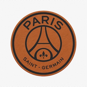 May include: A circular tan leather patch featuring a black printed logo. The design includes the word PARIS arched at the top, SAINT-GERMAIN at the bottom, and a central graphic of the Eiffel Tower silhouette above a small fleur-de-lis symbol.