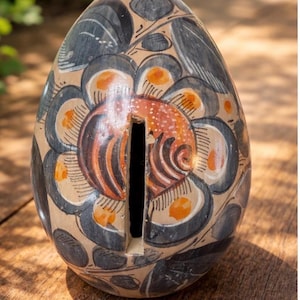May include: Hand-painted ceramic egg-shaped piggy bank. The bank features a floral design in black, orange, and cream colors. A vertical slot is cut into the top for inserting coins. The bank is sitting on a wooden surface.