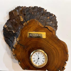 May include: A wooden clock featuring a gold-coloured clock face with Roman numerals. The clock is set in a piece of natural wood with dark brown, textured bark and a gold plaque that reads "AUSTRALIA".