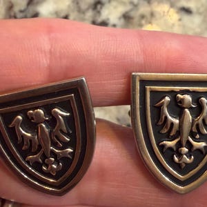 Puede incluir: Un par de gemelos metálicos, cada uno con forma de escudo con un emblema de águila plateado en relieve sobre un fondo negro hundido. Los bordes del escudo tienen un acabado en tono plateado pulido, creando un contraste marcado.