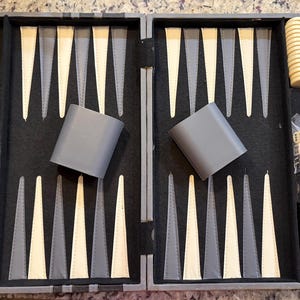 May include: Open backgammon set with a gray exterior and black interior. The game board features alternating white, gray, and black triangles. Includes gray dice cups, dice, and black game pieces. A handle and clasp are visible.