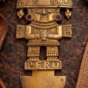 Puede incluir: Figura de metal dorado de una figura estilizada con un gran tocado y cuerpo rectangular. La figura tiene detalles morados y la palabra "PERU" está en la base. La figura está sobre una superficie marrón.