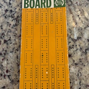 May include: A rectangular yellow Pleasantime continuous track two lane cribbage board with green and white text. The board features vertical columns of peg holes and a graphic of a peg at the bottom. The text reads Pleasantime, Continuous Track, Two Lane, Cribbage Board.