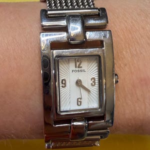 May include: A Fossil brand wristwatch with a silver-tone metal band and rectangular face. The watch face is white with black numerals at the 12, 3, 6, and 9 o'clock positions. The watch band is a woven chain style.