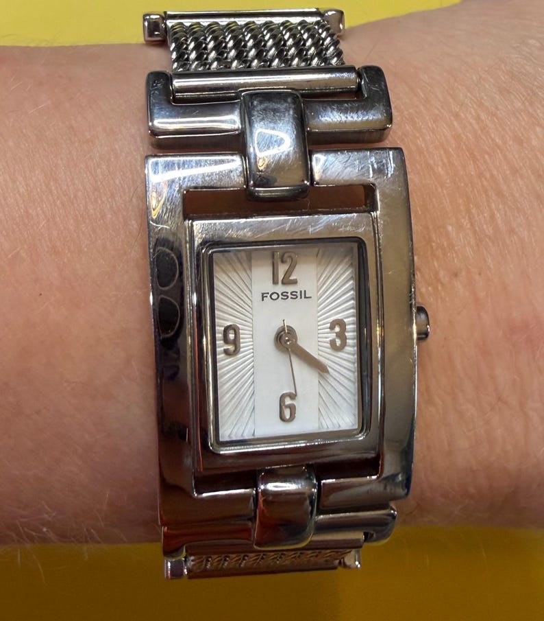 May include: A Fossil brand wristwatch with a silver-tone metal band and rectangular face. The watch face is white with black numerals at the 12, 3, 6, and 9 o'clock positions. The watch band is a woven chain style.