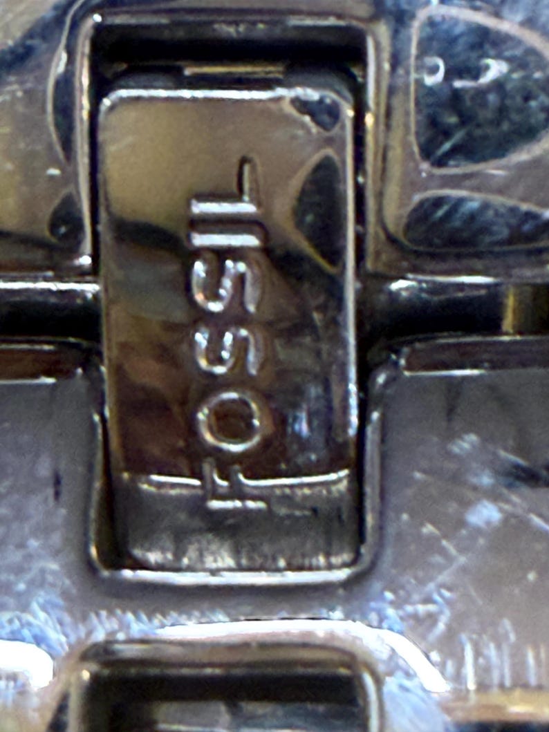 May include: Close-up of a silver watch clasp with the brand name "FOSSIL" and the number "15501" engraved on it. The clasp is rectangular and has a shiny, reflective surface.