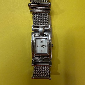May include: A silver Fossil watch with a rectangular face and a mesh band. The watch face is white with black numerals and hands. The watch is displayed against a yellow background.