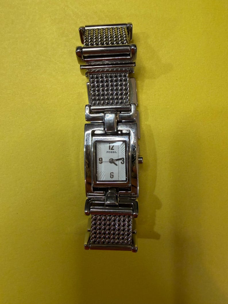 May include: A silver Fossil watch with a rectangular face and a mesh band. The watch face is white with black numerals and hands. The watch is displayed against a yellow background.
