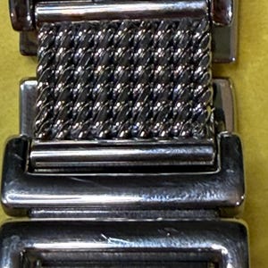 May include: Close-up of a silver-toned watch band. The band features a woven mesh center section and rectangular end pieces. The metal has a polished, reflective finish, and the design is simple and elegant.