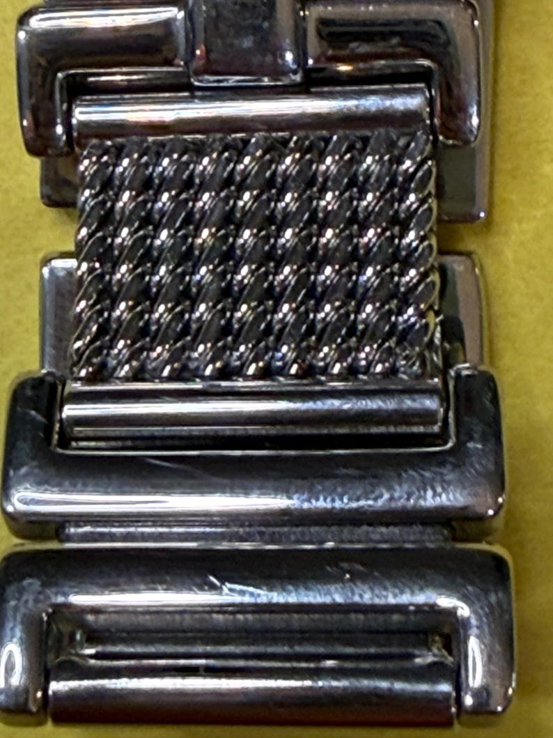 May include: Close-up of a silver-toned watch band. The band features a woven mesh center section and rectangular end pieces. The metal has a polished, reflective finish, and the design is simple and elegant.