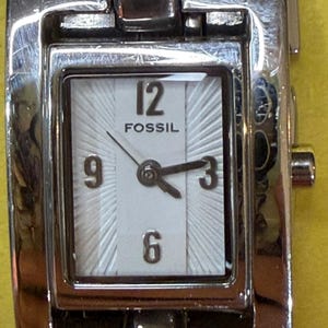 May include: A Fossil brand wristwatch with a silver-toned metal band and case. The watch face is square with a white background, black numerals, and hands. The watch displays the time as approximately 2:40.