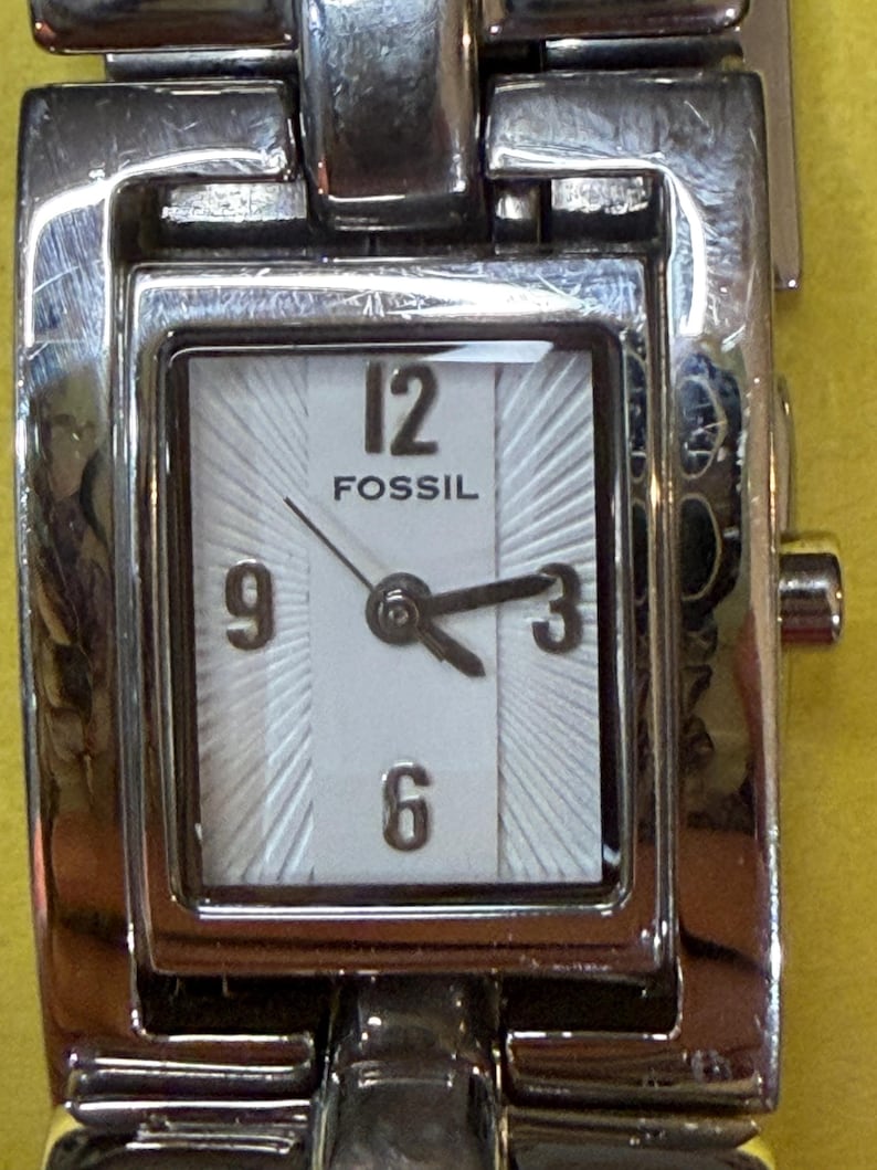 May include: A Fossil brand wristwatch with a silver-toned metal band and case. The watch face is square with a white background, black numerals, and hands. The watch displays the time as approximately 2:40.