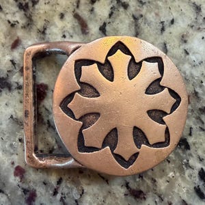 May include: A copper-colored belt buckle with a circular design featuring a stylized eight-pointed star. The buckle has a rectangular frame and a textured surface, suggesting a vintage or handcrafted aesthetic.
