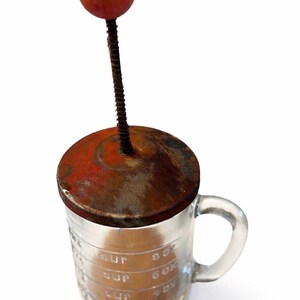 Könnte beinhalten: Vintage Messbecher aus Glas mit Griff, mit einem rostigen Metalldeckel und einer roten Holzkugel oben. Der Becher hat Messungen in Unzen und Tassen, perfekt für die Küche.
